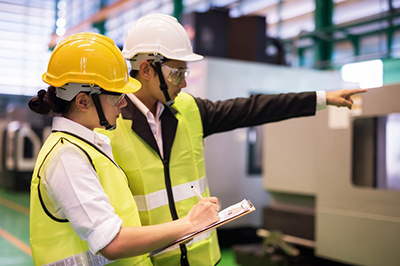 audit Deux ingénieurs en gilets jaunes et casques de sécurité effectuent une inspection sur un site industriel, l’un d’eux prenant des notes tandis que l’autre désigne une zone précise.
