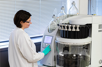 Chercheuse en blouse blanche manipulant un automate d’extraction dans un laboratoire d’analyse physico-chimique, avec des flacons en verre et un écran de contrôle.