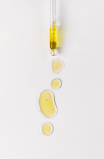 Pipette déposant des gouttes d’huile végétale jaune sur une surface blanche, illustrant une formulation cosmétique ou analytique.