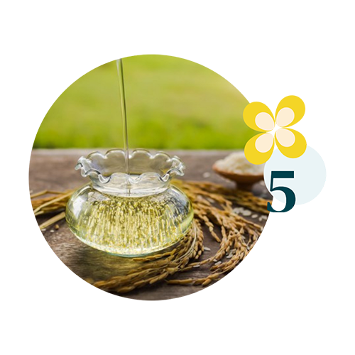 valeurs 5 Huile végétale dorée versée dans une carafe en verre posée sur une table en bois, entourée d’épis de riz, avec un décor naturel en arrière-plan et un motif floral accompagné du chiffre 5.